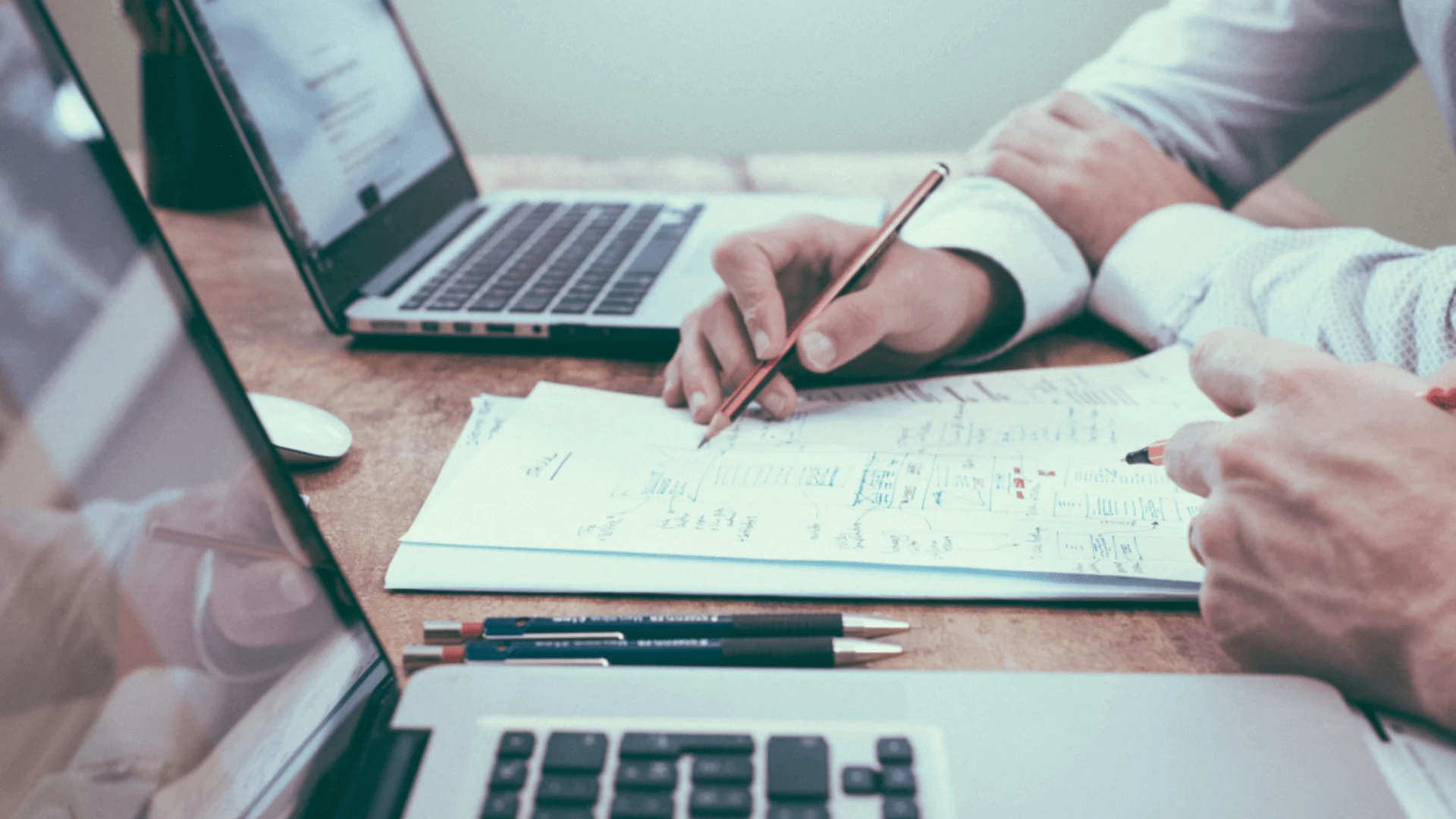 Two people review HR audit documents, taking notes beside open laptops on a desk.